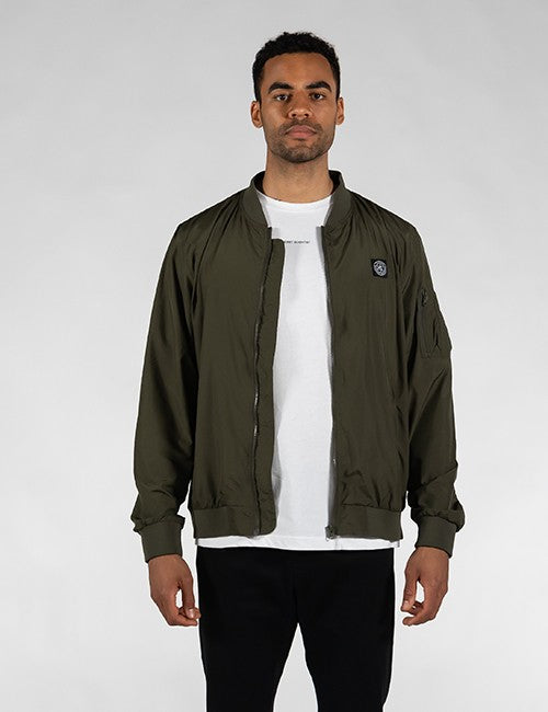 A man standing against a plain gray background wearing an open olive green bomber jacket over a plain white t-shirt and black pants.