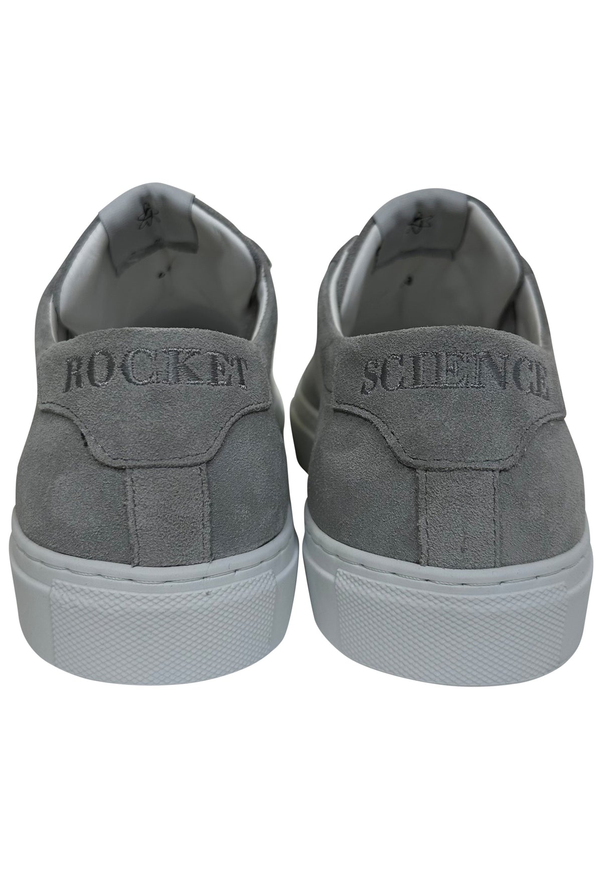 Heel view of a pair of low-top grey suede sneakers with white rubber soles.The left shoe has the word "ROCKET" embroidered in a dark grey font across the top of its heel tab, while the right shoe features the word "SCIENCE" embroidered in the same style.