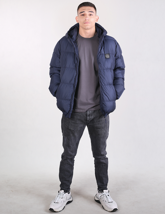 A man modeling a navy blue puffer jacket, worn over a dark gray t-shirt, dark gray jeans, and white sneakers, standing against a light gray studio background.