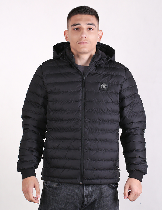 A medium-shot photograph of a young man with a short haircut, wearing a black quilted bubble puffer jacket with a hood and zipped up to the neck. The jacket has an elasticated cuff at the wrist and a circular logo badge on the left chest. He is facing forward against a plain white background, wearing dark gray jeans.