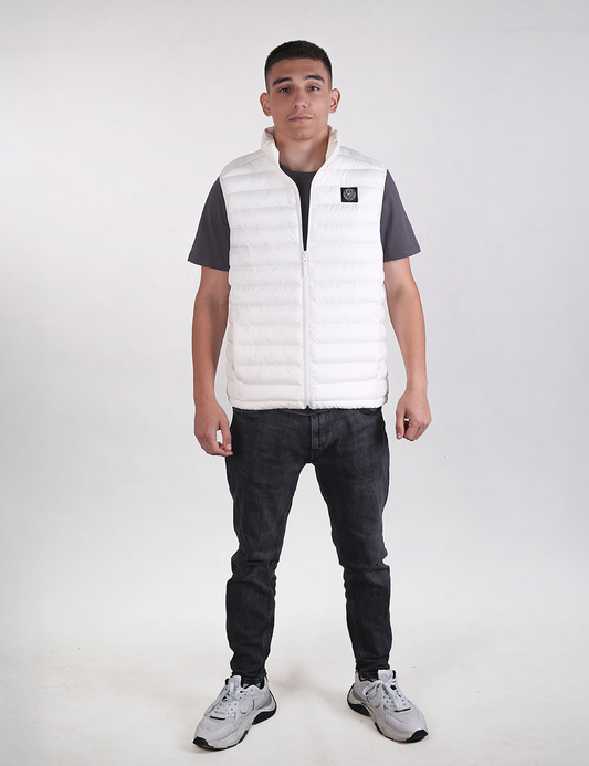 A man modeling a white puffer vest over a dark gray t-shirt, black jeans, and gray sneakers, standing against a light gray studio background.