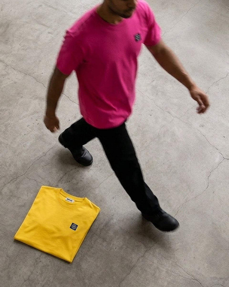 A man wearing a bright pink t-shirt and black pants walks across a concrete floor, with a folded yellow t-shirt placed on the ground nearby.