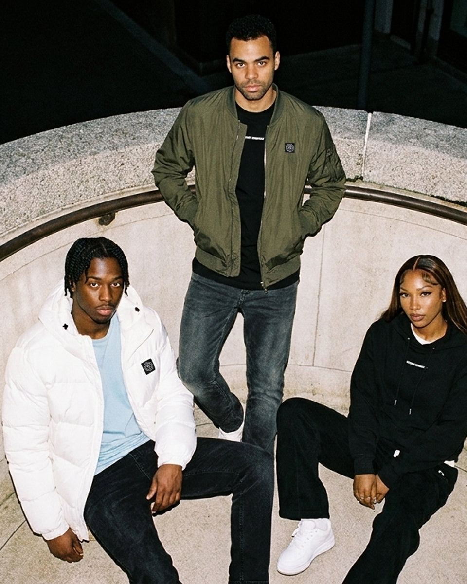Three models posing together outdoors at night in front of a concrete curved wall. The standing model wears an olive green bomber jacket over a black shirt and dark jeans.