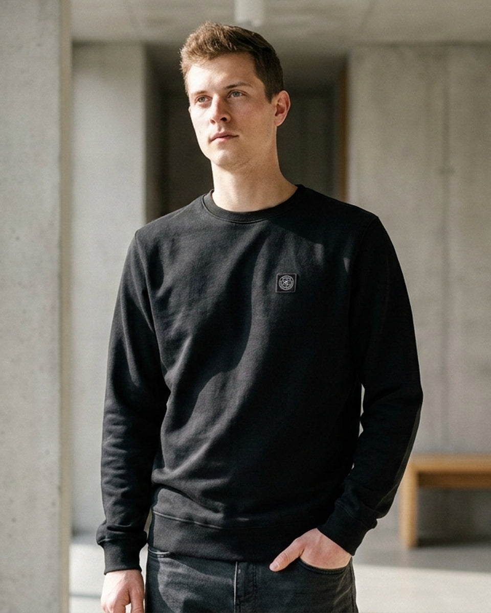 A man wearing a black crewneck sweatshirt with a small square logo patch on the left chest, standing in a modern concrete hallway.