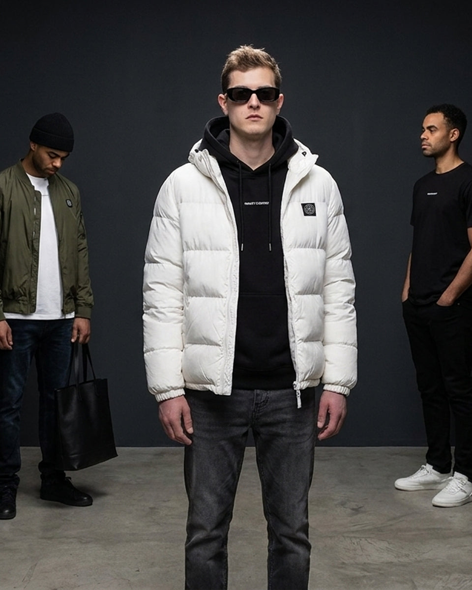 A model in the center wears a white puffer jacket over a black hoodie and dark jeans, standing in a studio with two other men in the background against a dark gray backdrop.