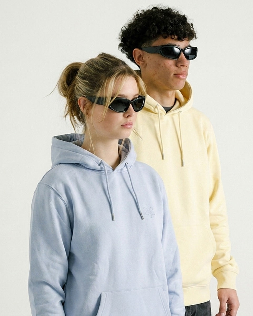 Two models wearing minimalist black sunglasses, with the model in the foreground wearing a light blue hoodie and the model behind wearing a pale yellow hoodie, set against a plain white background.