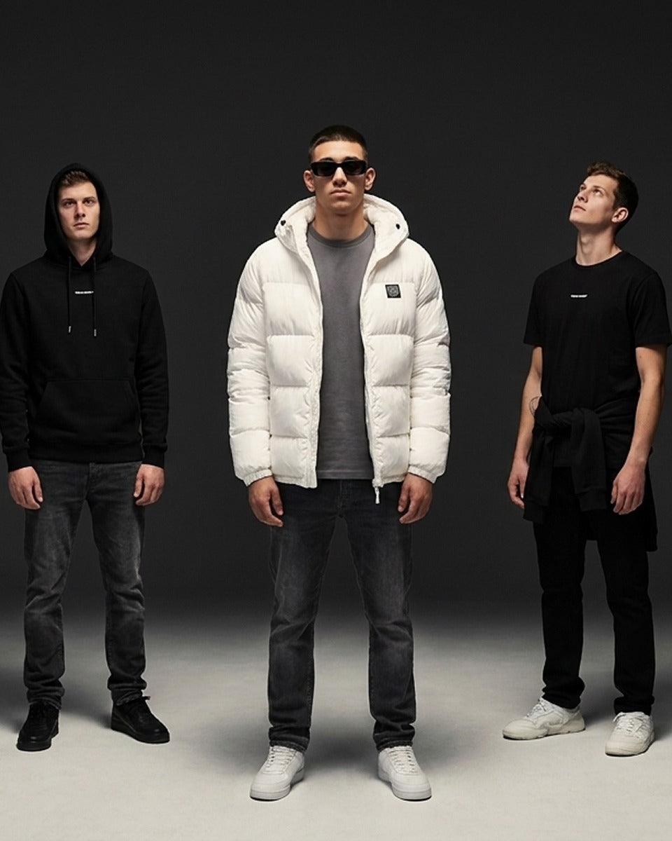 Three men standing against a dark studio background, each wearing a minimalist black apparel item: a hoodie, a puffer jacket, and a short-sleeve top. The central man wears a white puffer jacket over a gray shirt, dark jeans, and white sneakers, while the others wear black clothing and white sneakers.