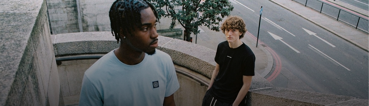 Two young men standing outdoors on a stone walkway next to a street. The man in the foreground wears a light blue t-shirt with a small black square logo on the chest, while the man behind him wears a black t-shirt.