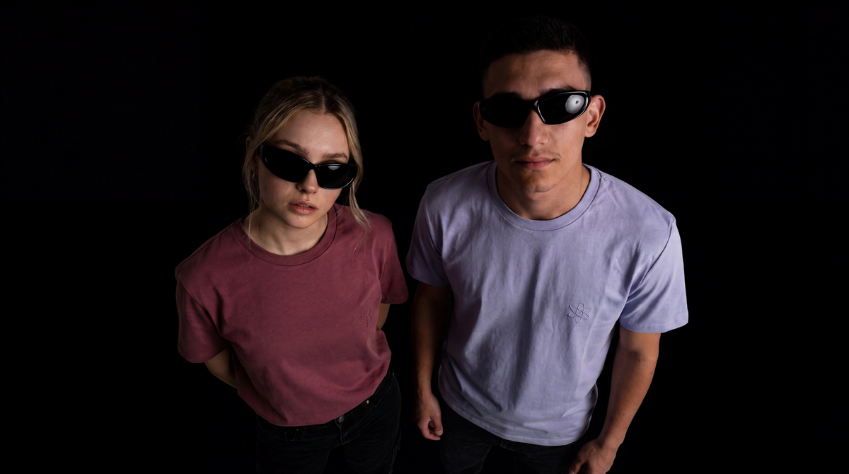 Two models wearing black oversized sunglasses, posed against a solid black background. The model on the left wears a muted rose pink t-shirt, and the model on the right wears a light lavender t-shirt.