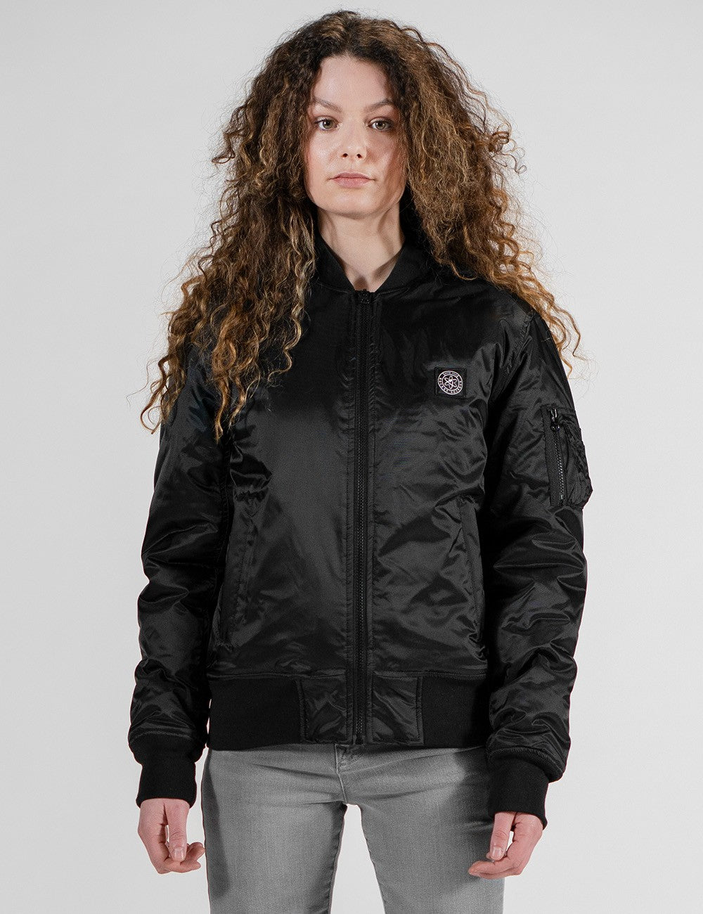 A woman wearing a black satin bomber jacket. The jacket has a small round logo patch on the chest and a zippered utility pocket on the upper left sleeve.
