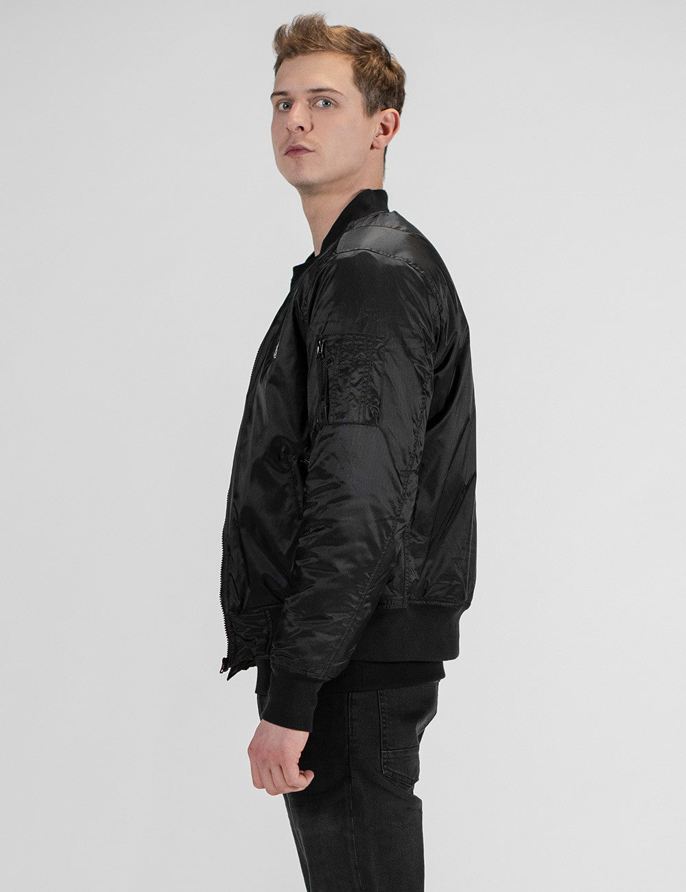A side-profile shot of a light-skinned young man with short, styled blonde hair looking toward the camera. He is wearing a black satin-finish bomber jacket featuring a zippered utility pocket on the upper sleeve, paired with black pants. 