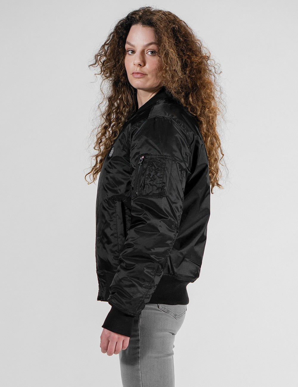A woman with long, curly, light brown hair stands in a side-profile view against a plain, light grey background. She is wearing a black nylon bomber jacket with ribbed cuffs, a ribbed waistband, and a zippered utility pocket visible on the upper left sleeve.