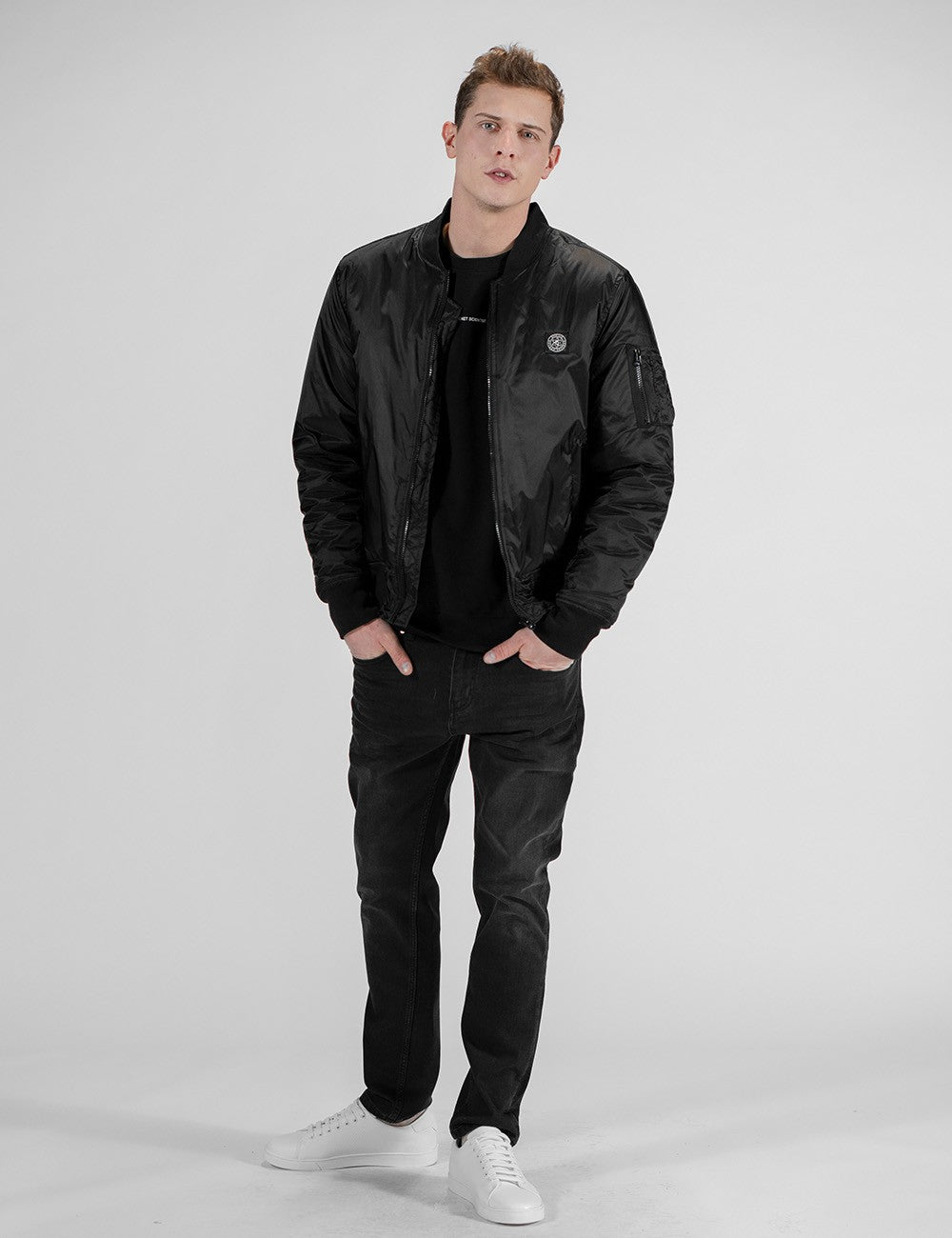 A man stands facing slightly to the right, looking forward. He wears a black bomber jacket zipped down, revealing a black crew neck shirt. The jacket has a small round patch on the left chest. He also wears dark-wash jeans and white sneakers.