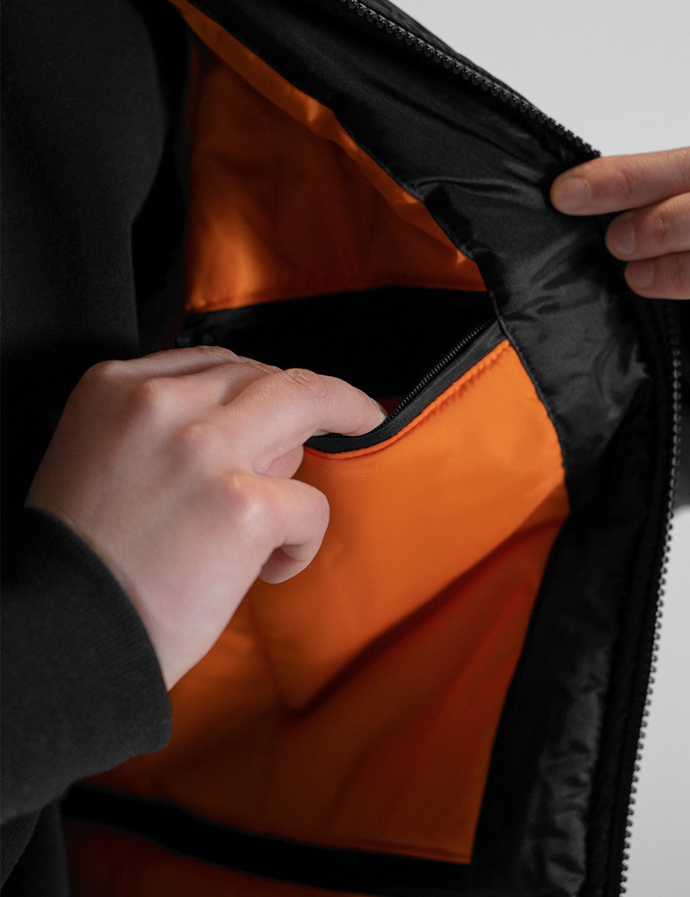 A close-up shot showing the bright orange interior lining of a black jacket. A person's left hand is reaching into an open internal pocket built into the orange fabric, while their right hand holds open the outer black zipper track.