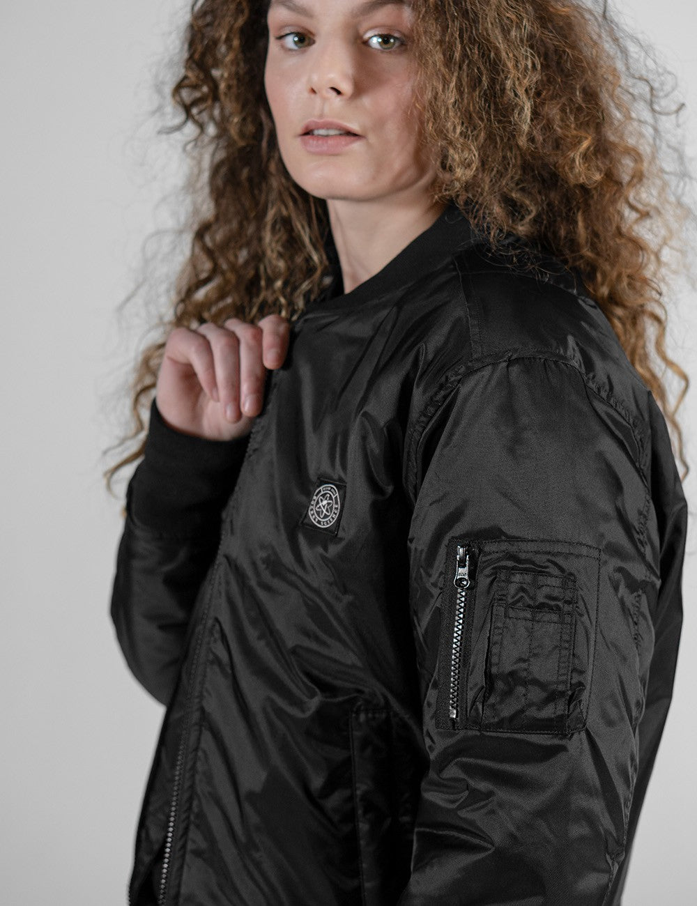 A close-up shot of a woman with long, curly brown hair wearing a shiny black bomber jacket against a light gray background. She is turned slightly to the side, looking directly at the camera. The jacket features a circular logo patch on the chest and a zipper pocket on the sleeve.