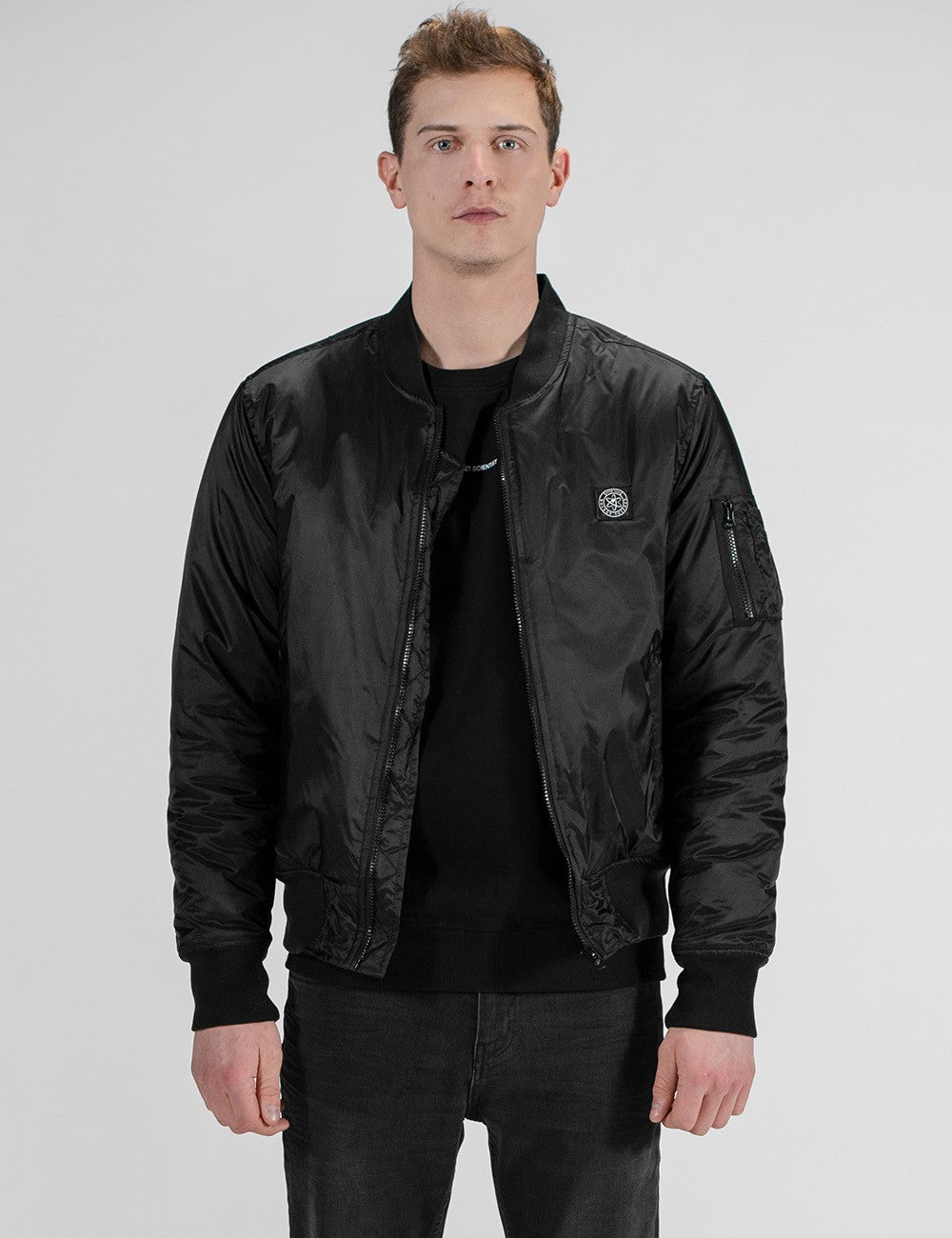 A male model with short brown hair stands facing forward against a plain white background, wearing a black satin bomber jacket over a black crew-neck t-shirt and dark gray jeans. The jacket features a ribbed baseball collar, elastic cuffs and hem, a zip sleeve pocket on the upper left arm, and a small circular logo patch on the left chest.
