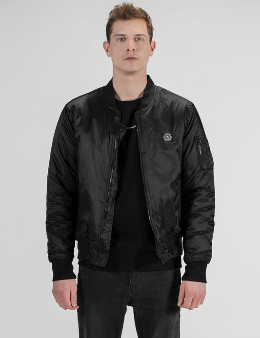 A male model with short brown hair stands facing forward against a plain white background, wearing a black satin bomber jacket over a black crew-neck t-shirt and dark gray jeans. The jacket features a ribbed baseball collar, elastic cuffs and hem, a zip sleeve pocket on the upper left arm, and a small circular logo patch on the left chest.