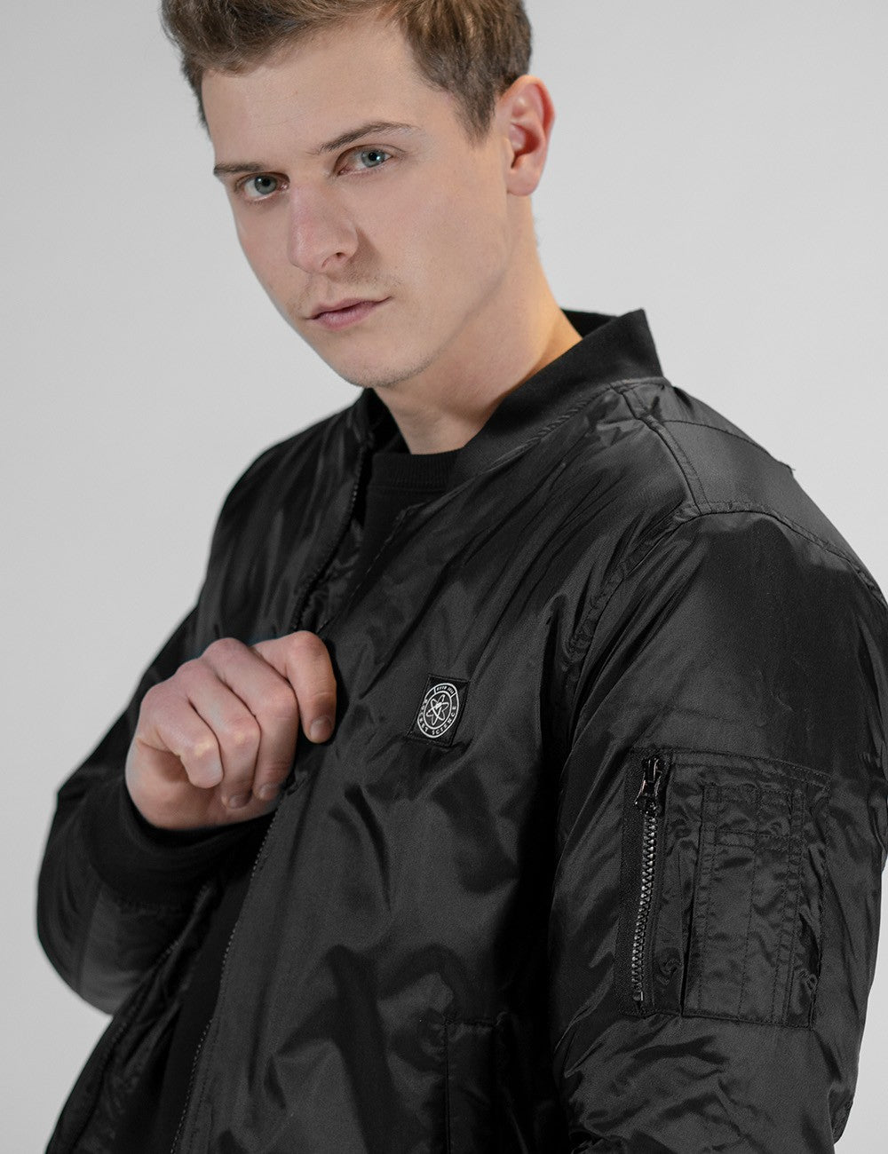 A man with short brown hair wearing a black Rocket Science bomber jacket. The jacket has a small round logo patch on the chest and a zippered utility pocket on the upper left sleeve.