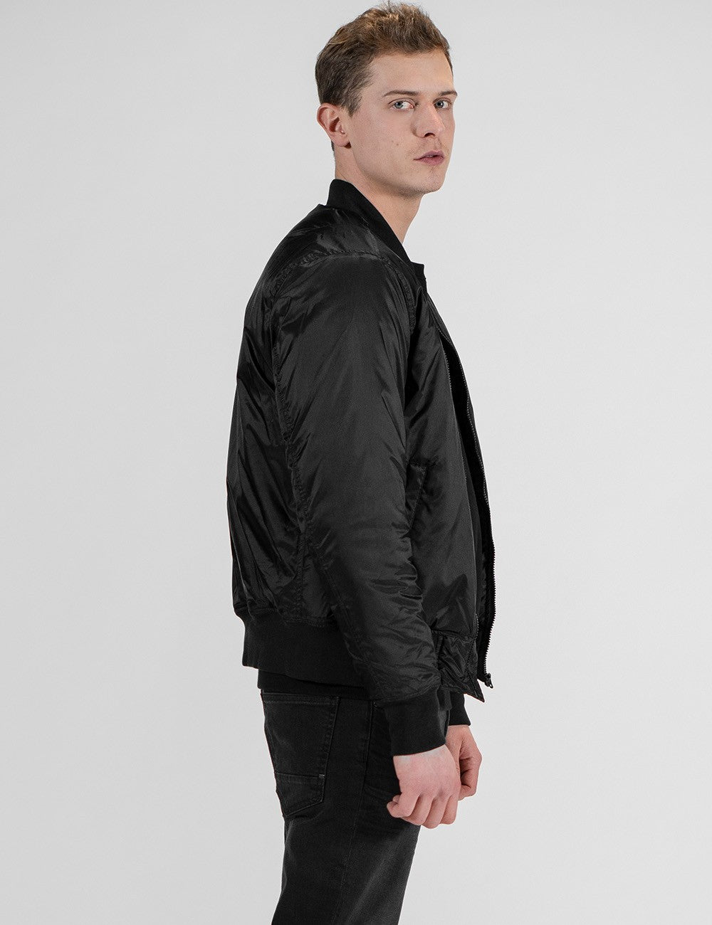 A young man with light brown hair stands in profile against a plain white background, wearing a black satin bomber jacket paired with black jeans.