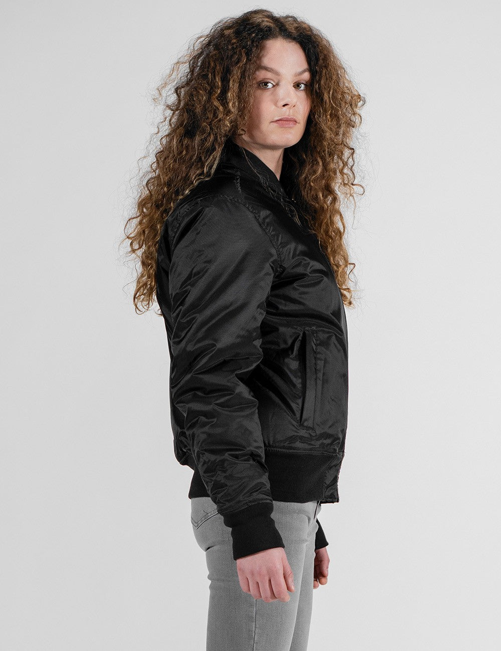 A woman with long, curly blonde and brown hair is seen from behind, wearing a black bomber jacket with ribbed cuffs and hem, paired with tight grey jeans .