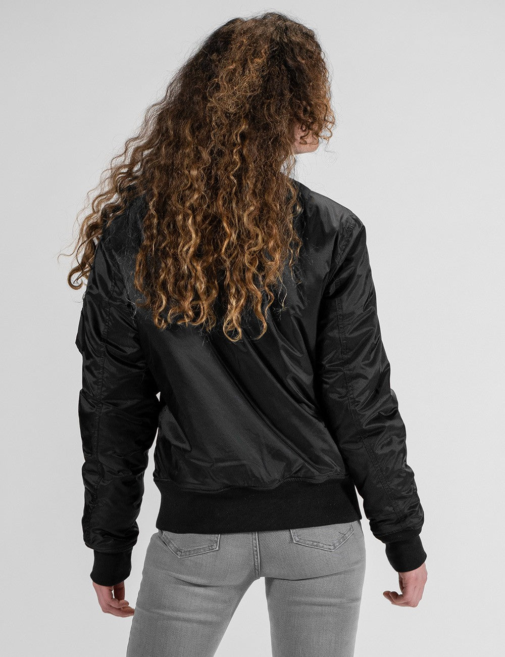 A female model with long, curly blonde and brown hair is seen from behind, wearing a black bomber jacket with ribbed cuffs and hem, paired with tight grey jeans.