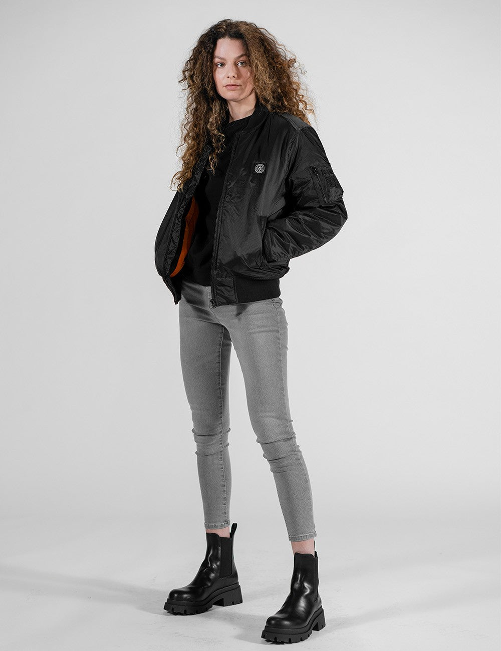 A full-length studio shot of a woman with long, curly brown hair facing forward, modeling a streetwear outfit. She is wearing a black Rocket Science bomber jacket with an orange lining, a black crewneck top underneath, tight grey skinny jeans, and chunky black leather lug-sole boots. 