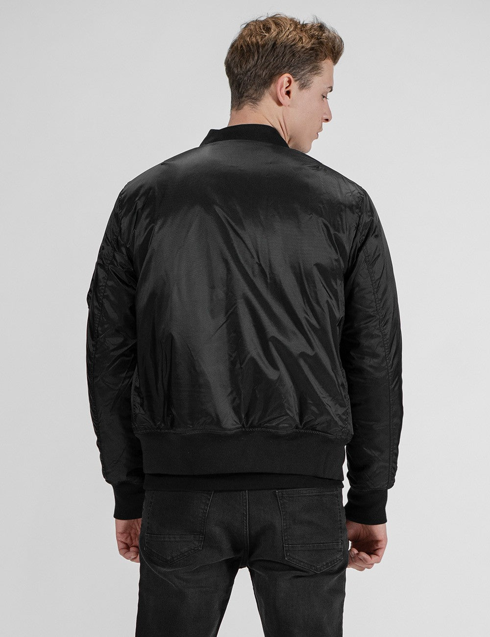A man viewed from behind wears a black bomber jacket over black jeans .