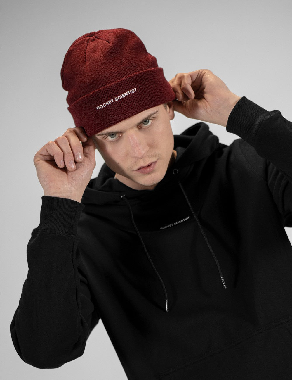 A man wearing a black hoodie and a dark red knit beanie with 'ROCKET SCIENTIST' embroidered in white on the cuff, posing against a neutral gray background.