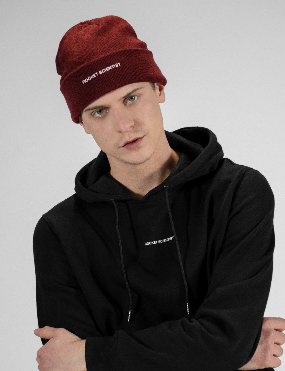 A man wearing a maroon knit beanie and a black hoodie, both with white embroidered 'ROCKET SCIENTIST' branding, posed against a light gray studio background.