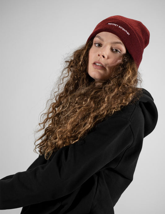 A model with long curly brown hair wearing a dark red beanie with 'ROCKET SCIENTIST' in white text and a black hoodie, photographed against a neutral gray background.