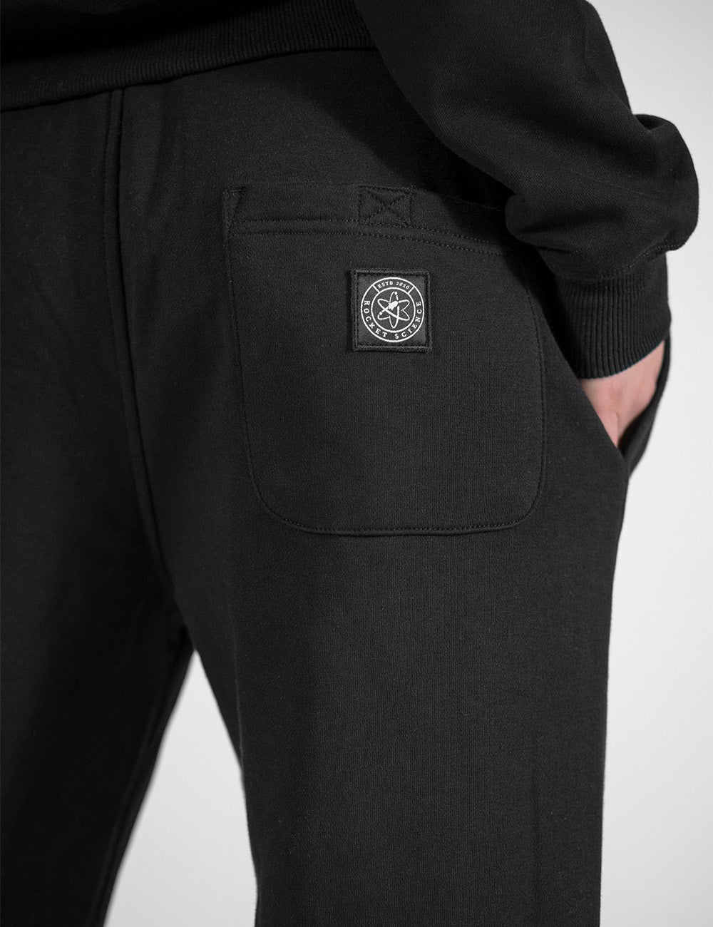 A close-up photograph focuses on the back pocket of a pair of black fabric sweatpants. Stitched onto the lower center of the pocket is a circular black patch featuring a white graphic outline of an atom surrounded by text that reads "ESTB 2012" and "ROCKET SCIENCE."