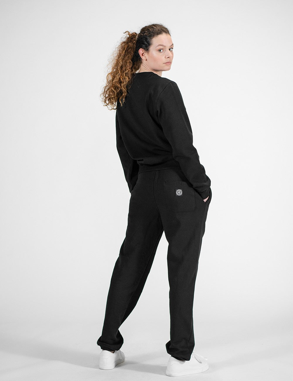A rear pose of a young woman with long, curly brown hair tied back in a ponytail stands facing away from the camera while looking back over their right shoulder. They are wearing an all-black casual outfit consisting of a long-sleeve sweatshirt and matching sweatpants with a small circular Rocket Science logo on the back pocket, paired with white sneaker.