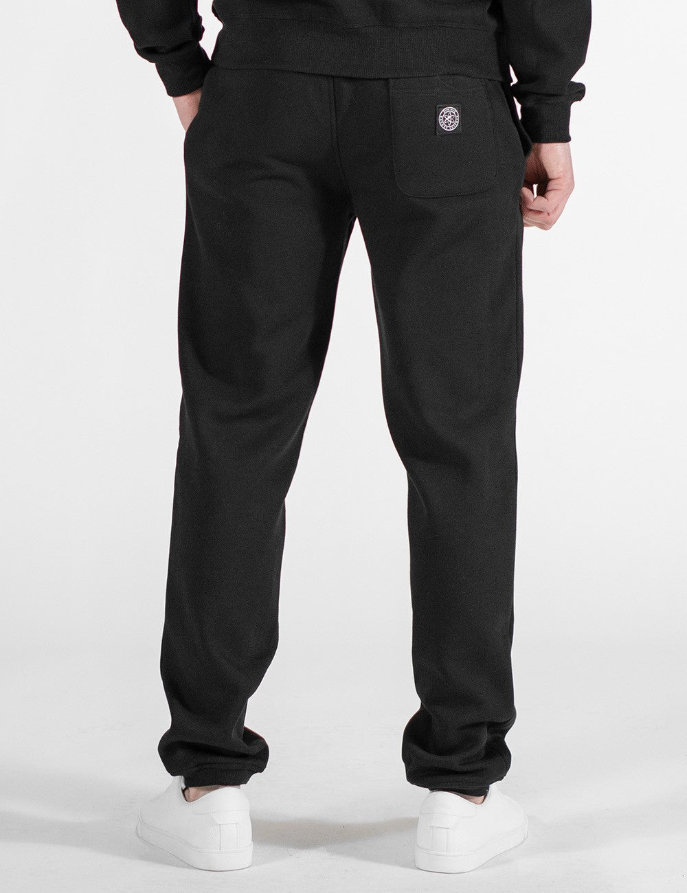 This image shows rear pose of a person wearing a casual black crew-neck sweatshirt and matching black sweatpants, paired with white sneakers.