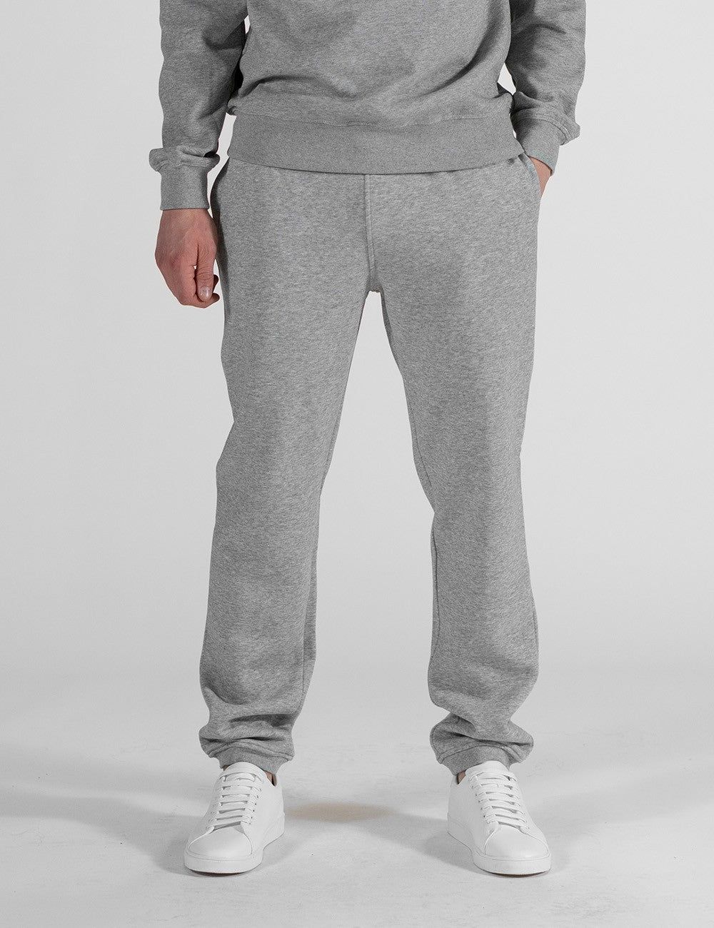 The image centers on a pair of heather grey sweatpants with a relaxed, straight-leg fit and classic elasticated ankle cuffs. They are styled as part of a matching loungewear set and paired with clean white leather sneakers.