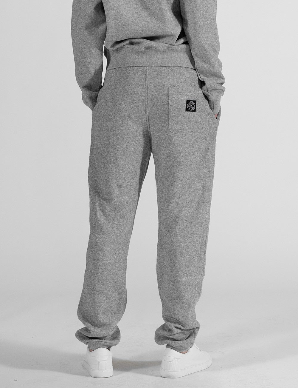 The image shows a rear pose of a pair of grey heather sweatpants with an elastic waistband, drawstrings, and a small square logo patch on the back pocket area.