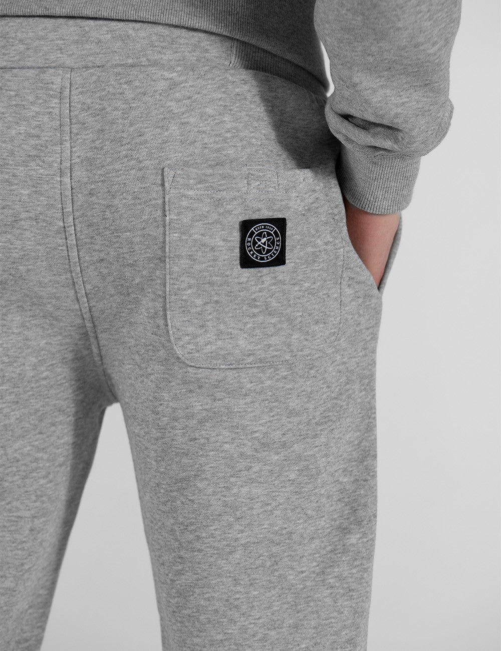 A close-up shot from behind shows a person wearing heather grey sweatpants with a back patch pocket featuring a black, circular Rocket Science logo patch.