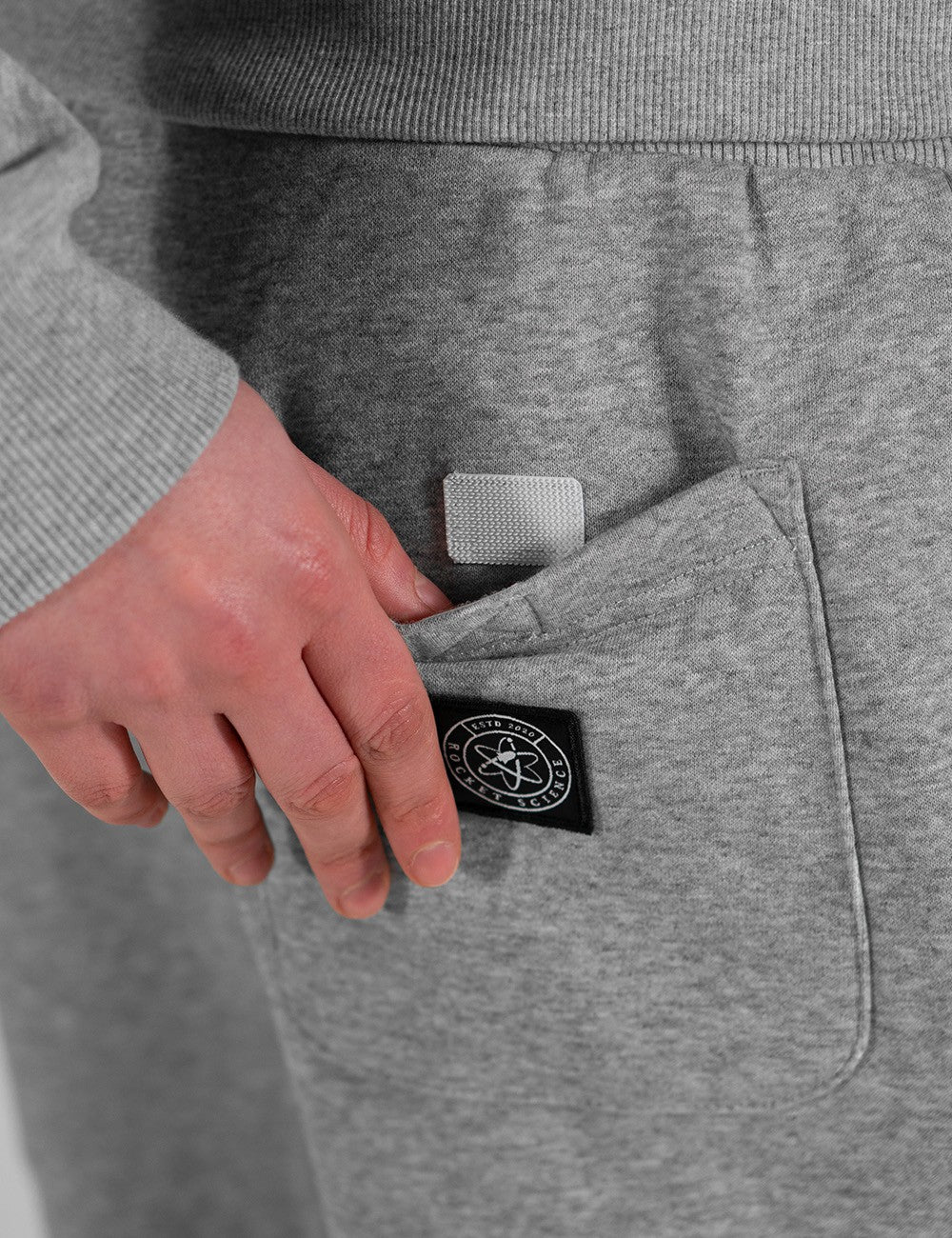 The image shows the Heavy Sweatpants, Heather Grey by Rocket Science, featuring a back pocket with a velcro fastener and a round logo patch.