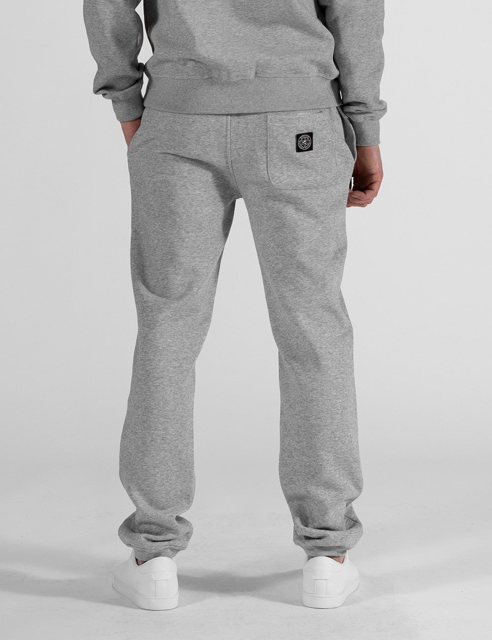 The image centers on a pair of heather grey sweatpants with a relaxed, straight-leg fit and classic elasticated ankle cuffs,  and a logo patch on the right back pocket. They are styled as part of a matching loungewear set and paired with clean white leather sneakers.