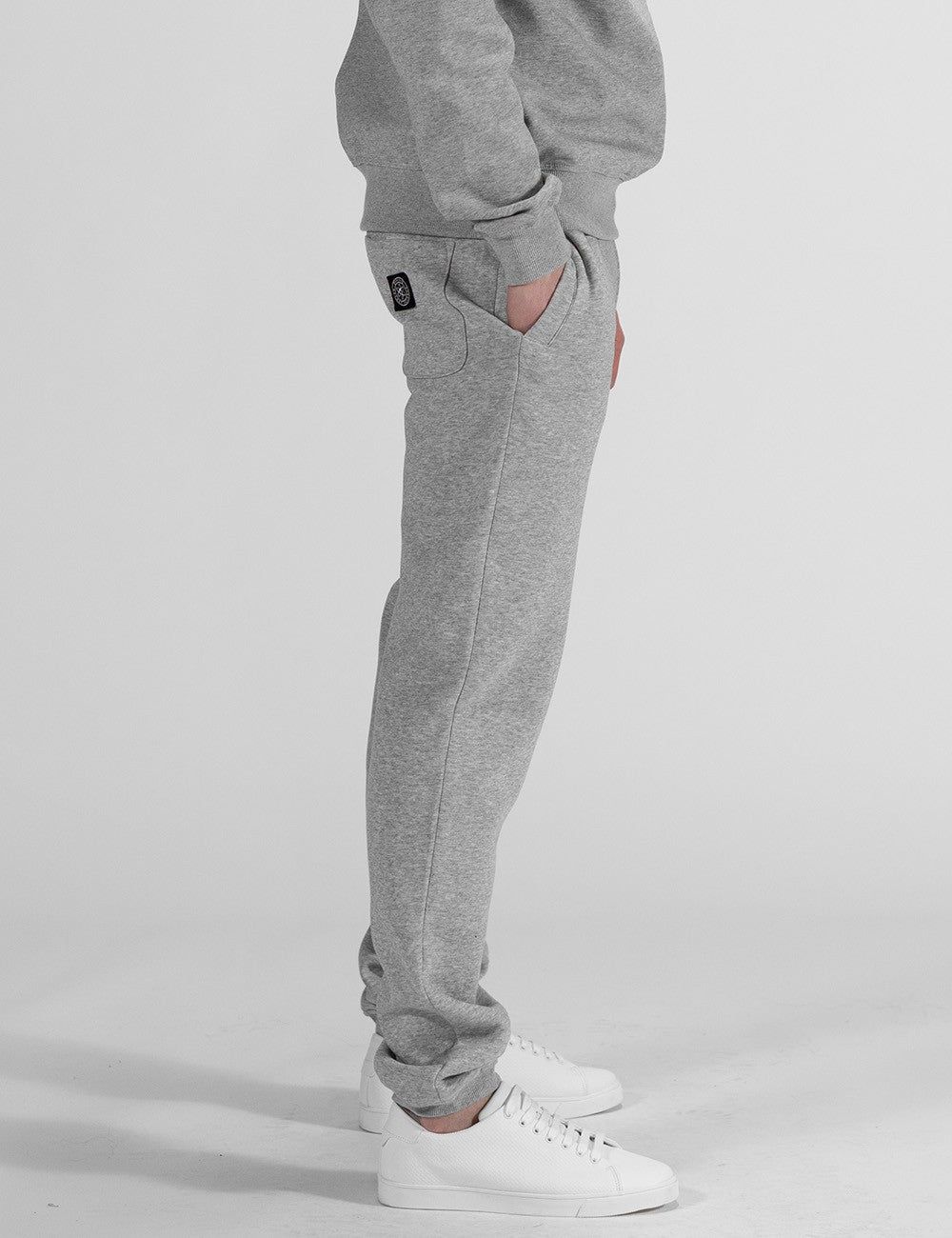 The image centers on a pair of heather grey sweatpants with a relaxed, straight-leg fit and classic elasticated ankle cuffs and a patch logo on the back pocket. They are styled as part of a matching loungewear set and paired with clean white leather sneakers.