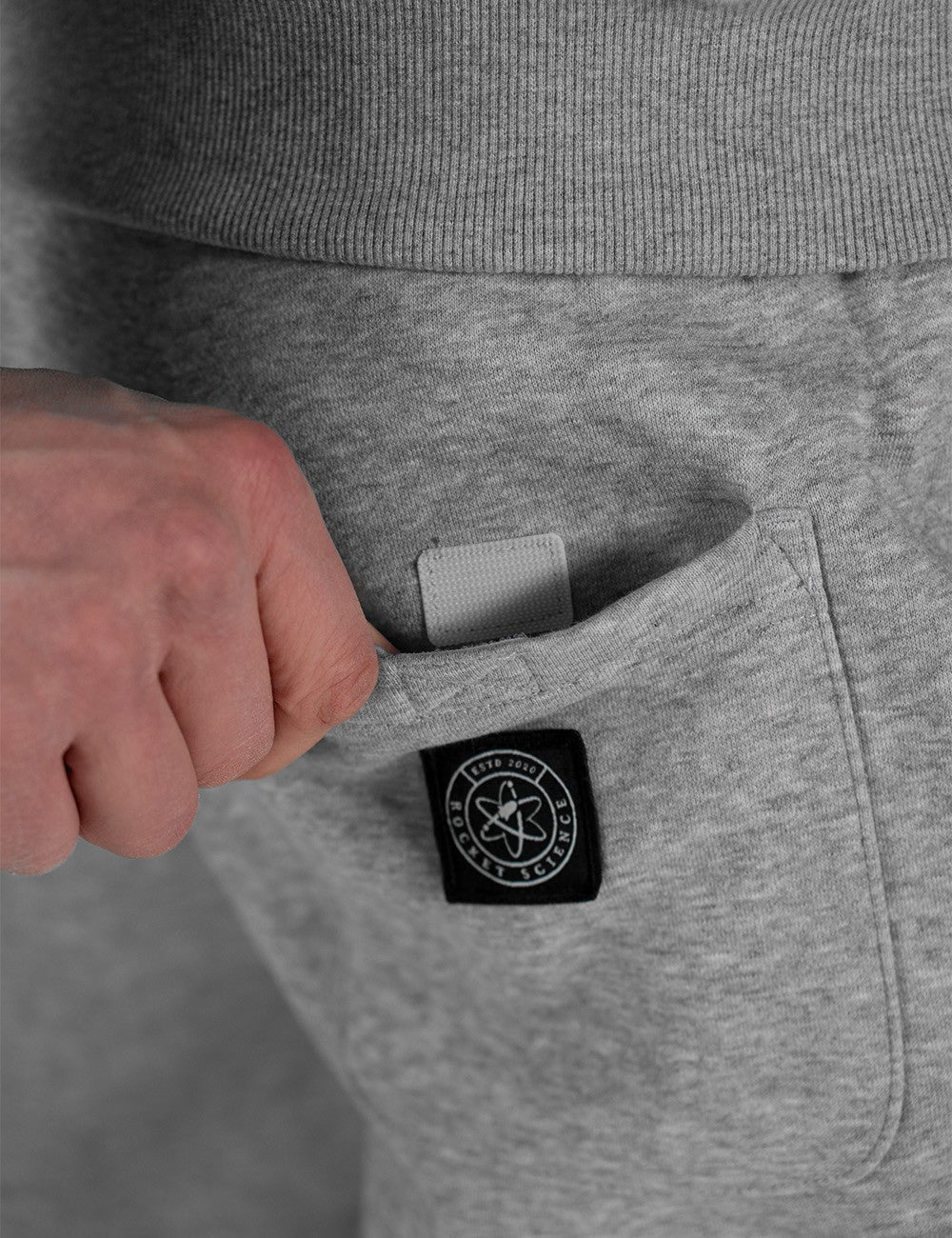 A close-up shot from behind shows a person wearing heather grey sweatpants with a back patch pocket featuring a black, circular Rocket Science logo patch.
