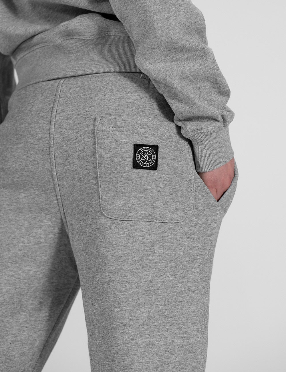 The image shows a pair of grey sweatpants featuring a square logo patch on the back pocket.