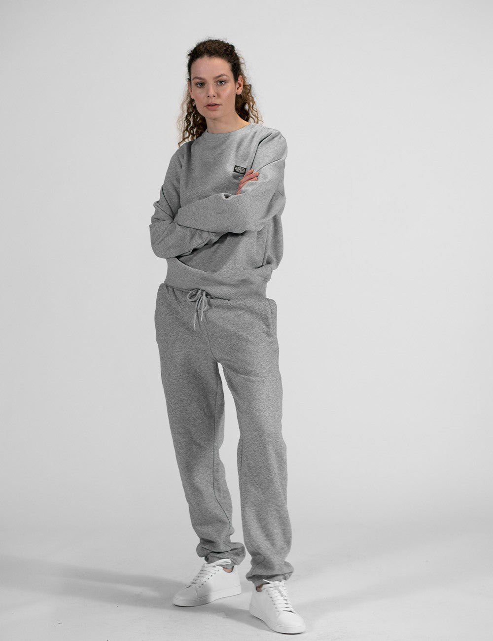 This image shows a young woman wearing  pair of casual grey sweatpants with an elastic drawstring, a crewneck sweatshirt , and white sneakers.
