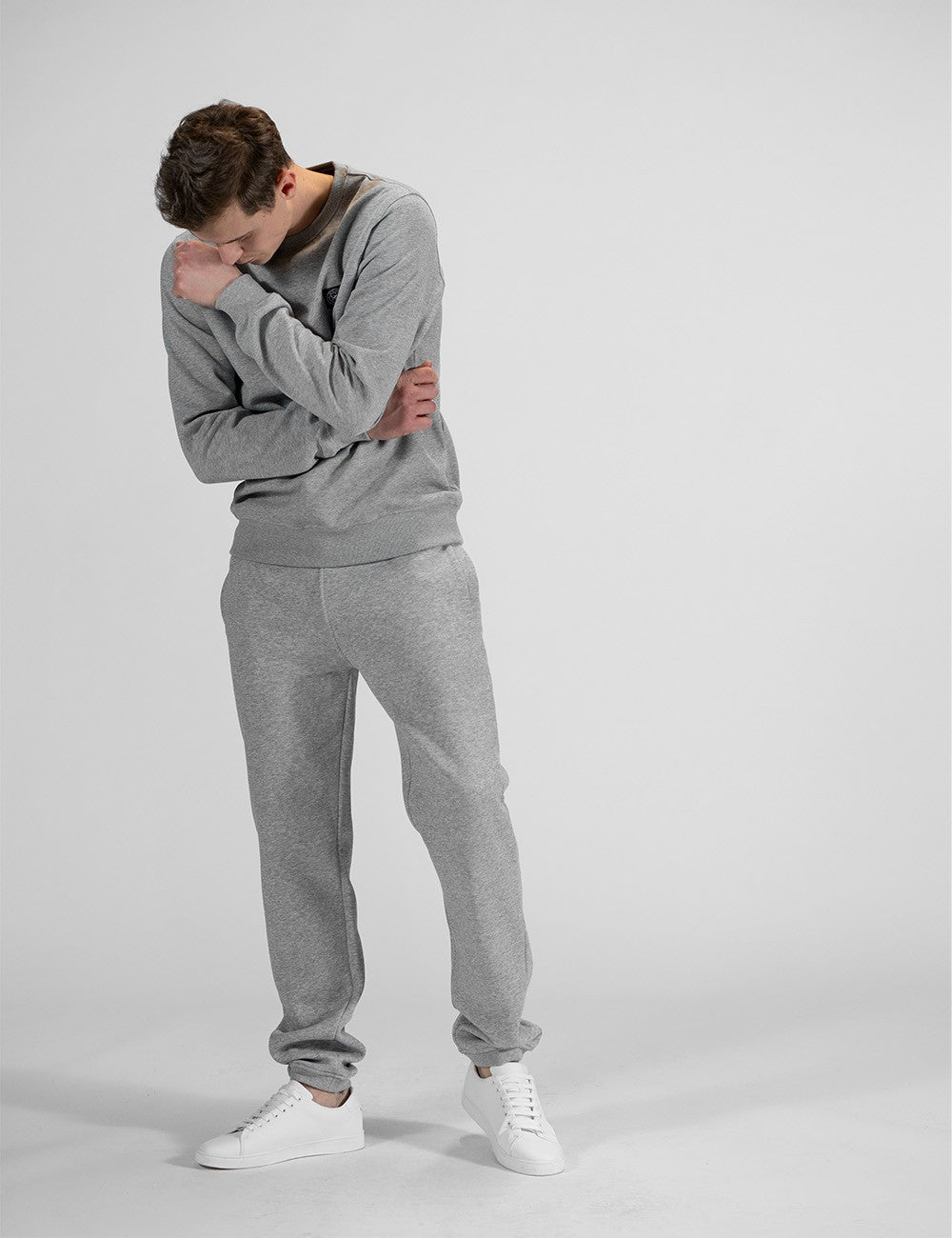 A full-body shot of a young man posing against a plain light grey background, showcasing a matching heather grey loungewear tracksuit by the brand Rocket Science. He stands angled slightly to the left, looking down thoughtfully over his shoulder with his hands resting relaxed across his chest and midsection.