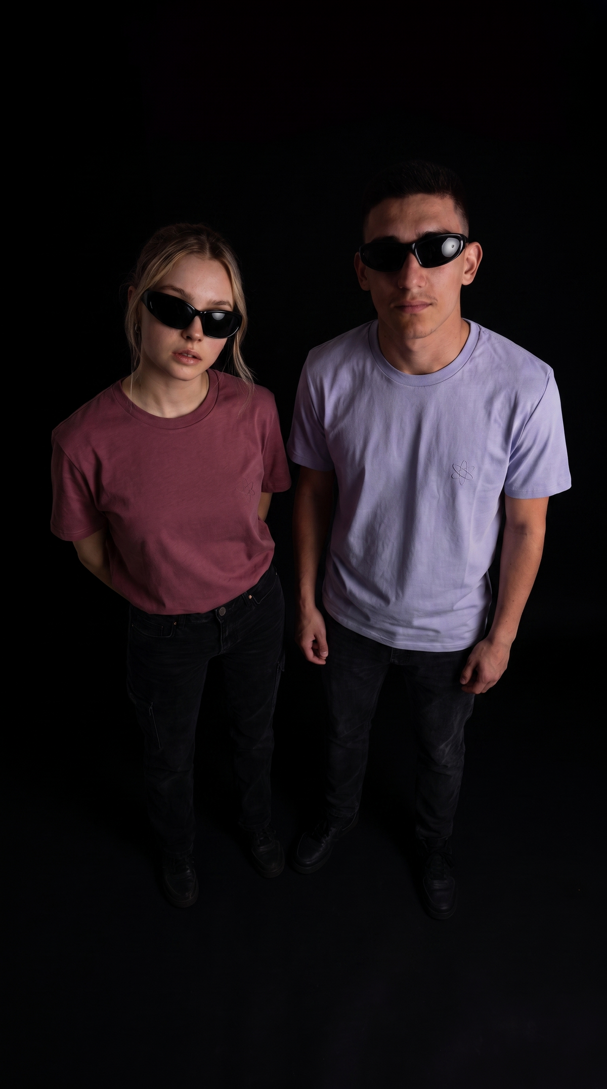 Two models wearing oversized black sunglasses and casual t-shirts, standing against a solid black background.