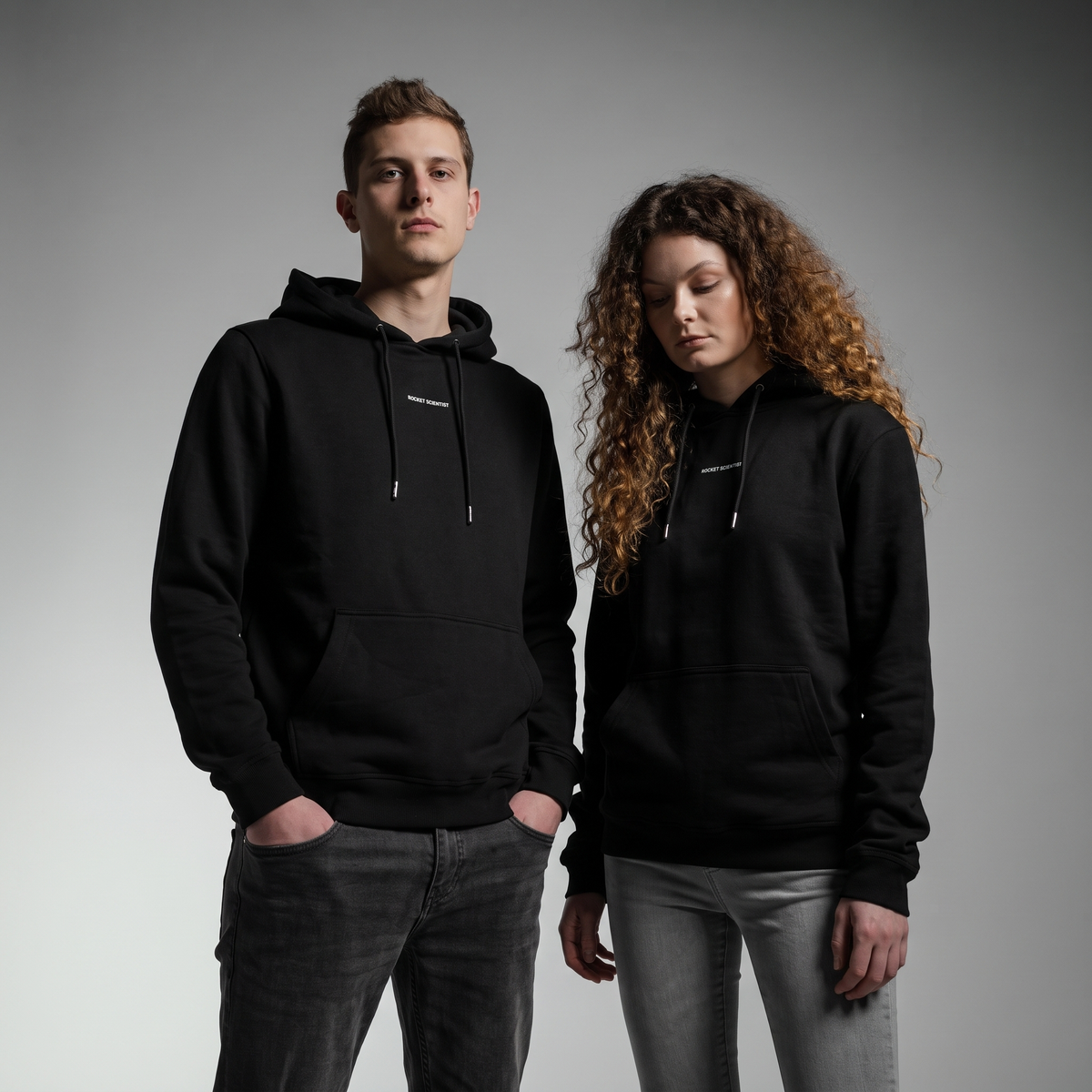 A man and a woman standing side-by-side wearing matching black pullover hoodies with white drawstrings and a small white text logo on the chest, photographed against a neutral gray studio background.
