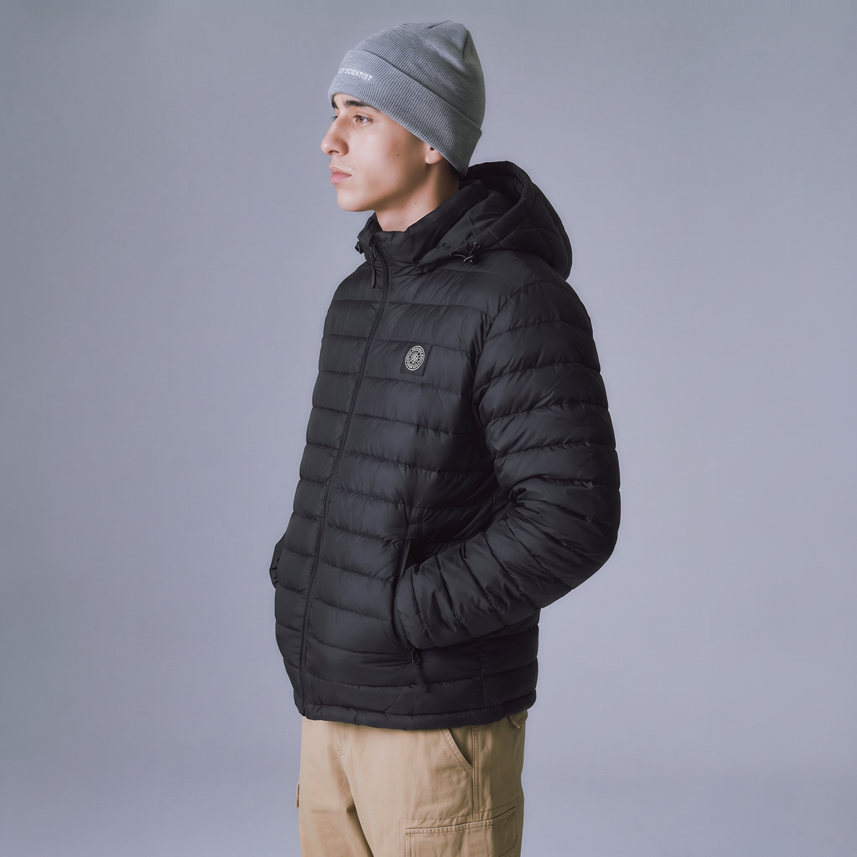 A man in profile wearing a black hooded puffer jacket, a gray knit beanie, and tan pants, photographed against a plain light gray studio background.
