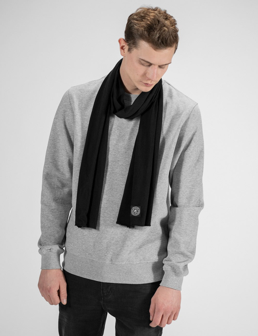 A close-up view highlighting a Black  jersey scarf from Rocket Science, loosely wrapped around a man's neck. The fabric hangs smoothly down the front of a grey heather sweatshirt. A prominent detail is the small, square dark logo patch stitched near the bottom edge of the scarf's right end.