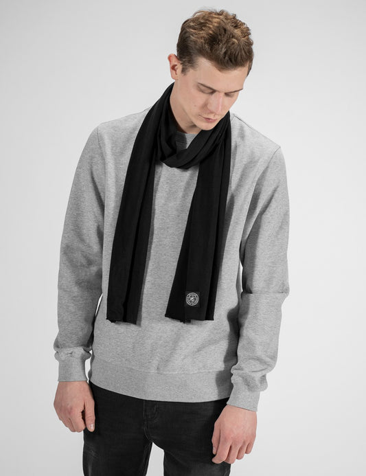 A close-up view highlighting a Black  jersey scarf from Rocket Science, loosely wrapped around a man's neck. The fabric hangs smoothly down the front of a grey heather sweatshirt. A prominent detail is the small, square dark logo patch stitched near the bottom edge of the scarf's right end.