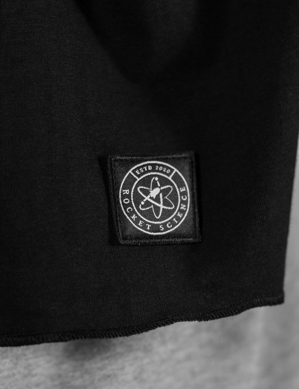 A close-up view highlighting a black jersey scarf from Rocket Science. A prominent detail is the small, square dark logo patch stitched near the bottom edge of the scarf's right end.
