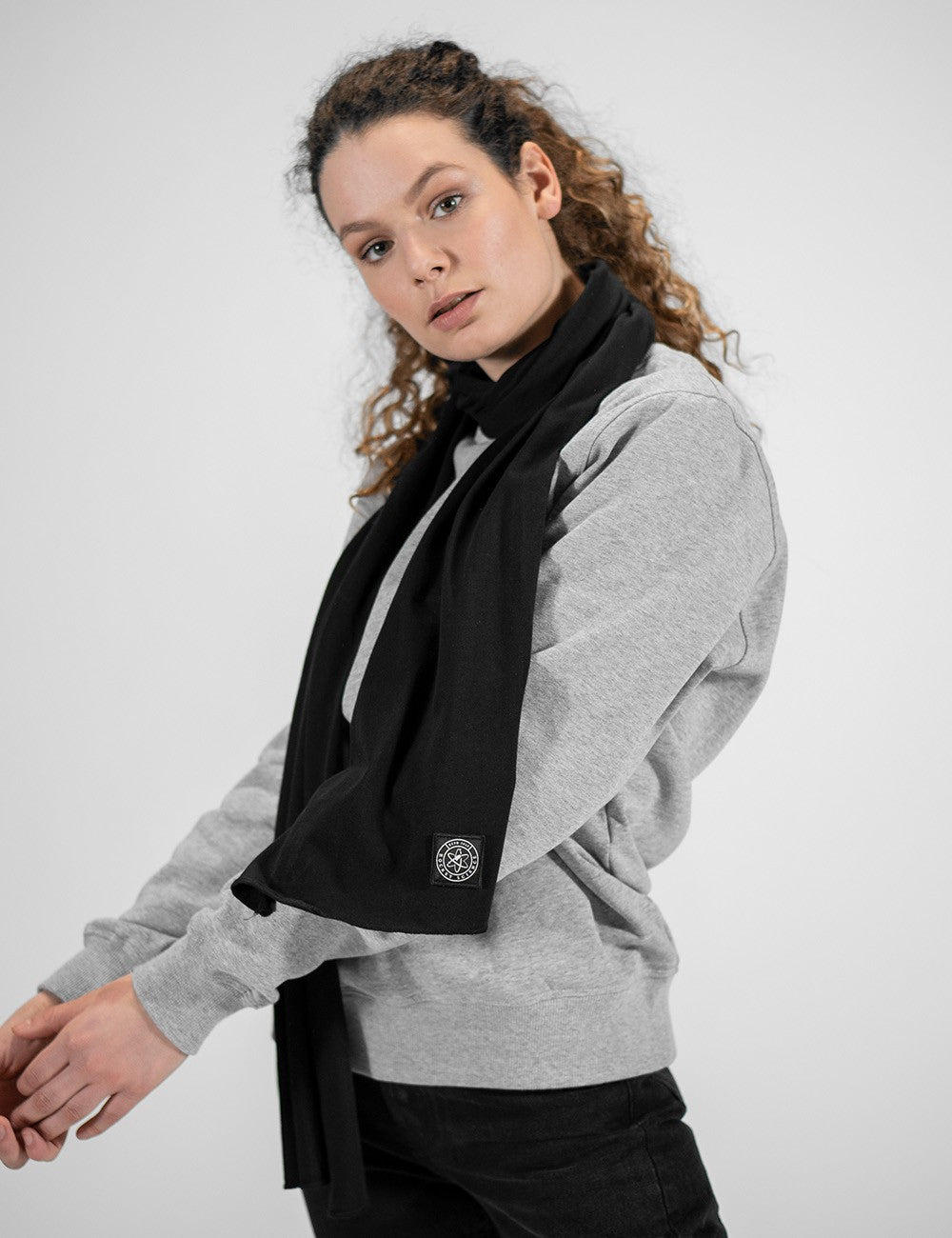 A close-up view highlighting an olive green jersey scarf from Rocket Science, loosely wrapped around a woman's neck. The fabric hangs smoothly down the front of a grey sweatshirt. A prominent detail is the small, square dark logo patch stitched near the bottom edge of the scarf's right end.